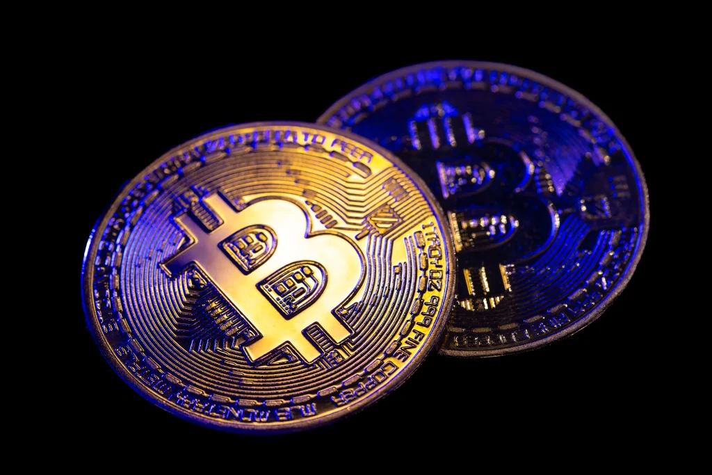 Two physical Bitcoin coins are displayed against a black background, one slightly overlapping the other, with gold and blue lighting reflecting off their surfaces.
