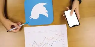A person holds a smartphone next to an inverted Twitter logo on paper. Below, a chart with fluctuating lines indicates data analysis on a wooden table.