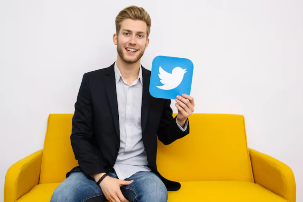 Young man in a black blazer sits on a yellow couch, smiling while holding a blue Twitter Influencers. The scene conveys a social media theme.